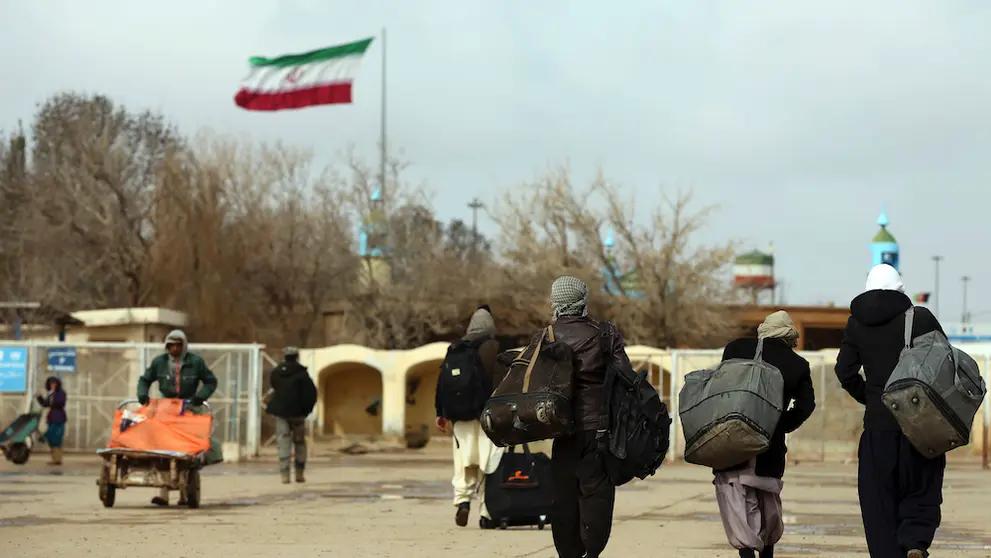 Afghan migrants walk away with their belongings as the Iranian flag flies in the background